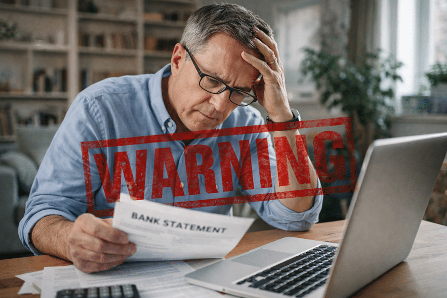 Man at a laptop holds a bank statement, with a red 'WARNING' overlay signaling financial alert or fraud risk.