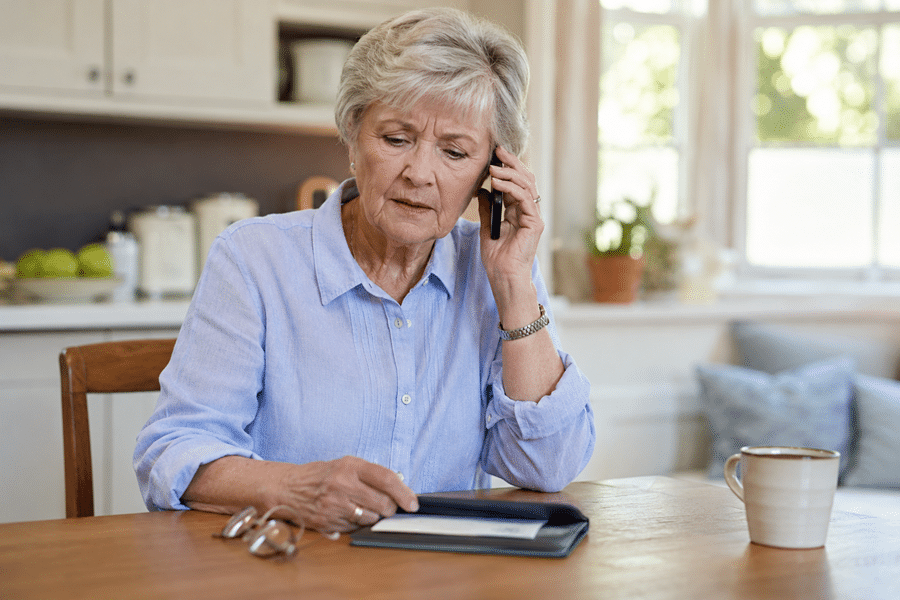 Senior woman in a blue shirt sits at a kitchen table on a cell phone, with a wallet and papers nearby and a mug on the table.